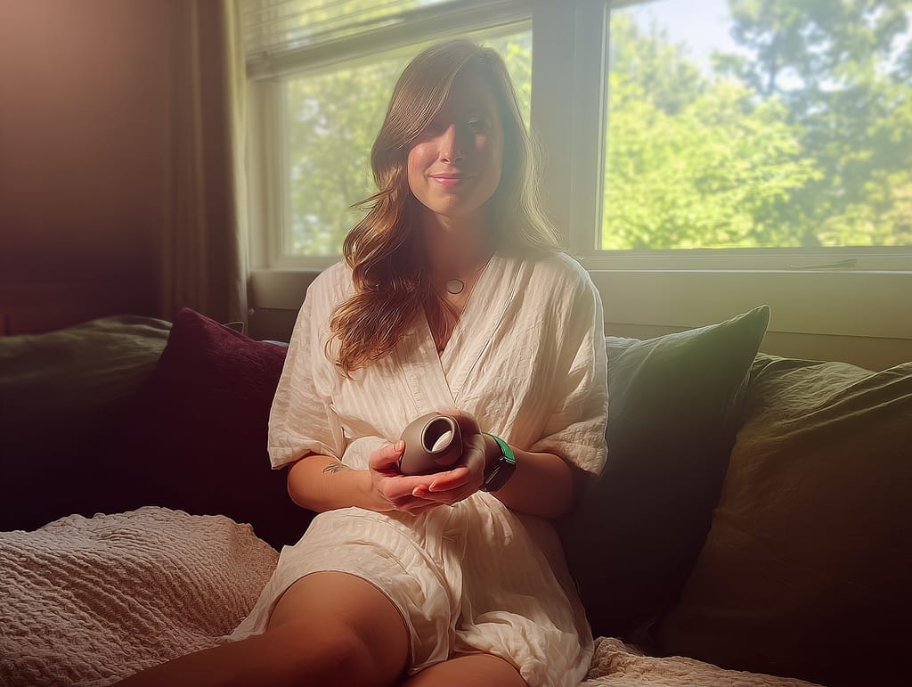   A warm, candid RAW photo taken from a distance, showing a young woman in a cozy, softly lit room, sitting comfortably on a couch or the edge of a bed. She holds a small, discreet, elongated personal wellness object in one hand smooth and matte, no longer than the span of two hands, with a soft circular opening at one end. The mood is light and relaxed. She’s smiling subtly or looks thoughtful in a natural, everyday way. The scene captures a quiet, pleasant moment at home, with warm daylight, cozy textures, and a peaceful vibe. Framed from farther away the woman is part of the environment, not the central focus.